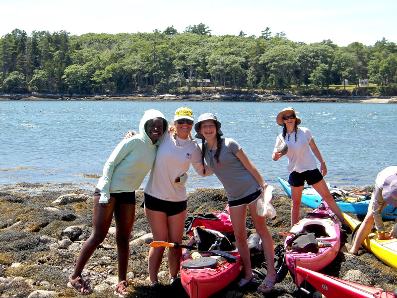 The image shows four young women on a rocky beach next to a body of water. Three of the women are standing next to kayaks, while one is further down the beach. The women appear to be enjoying a sunny day outdoors, possibly after a kayaking trip. The background features a treeline and the calm water of the bay.